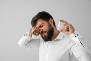 homme montrant des gouttes pour les yeux et se frottant un oeil. Young man with eye drops feeling discomfort on grey background.
