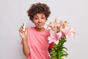 Femme avec un haut rose et les yeux rouges, au cheveux bouclés tenant des fleurs de lys et des gouttes pour les yeux